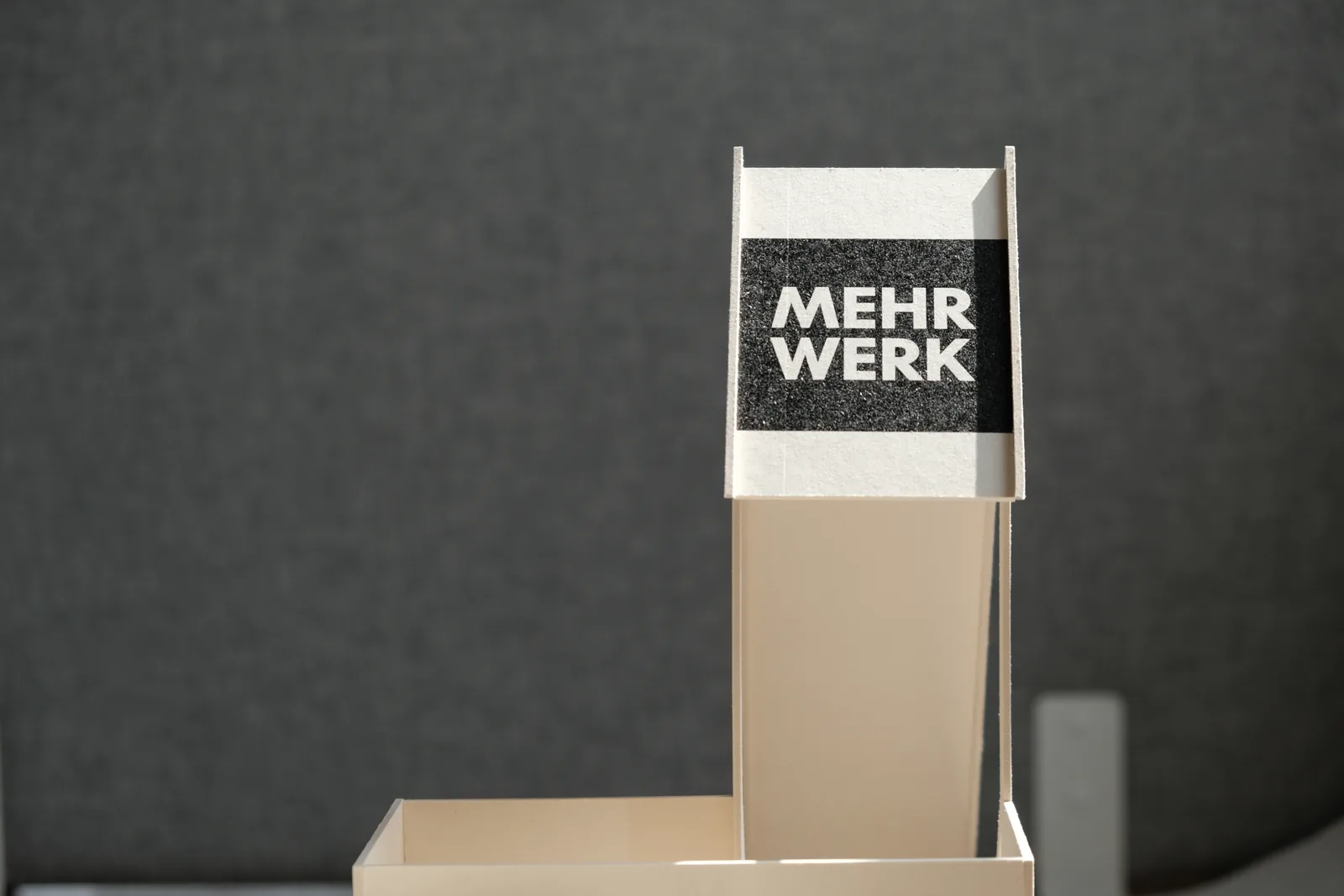 Modellfoto: der geschlossene Bau vor grauem Grund, oben die Tafel MEHR WERK.